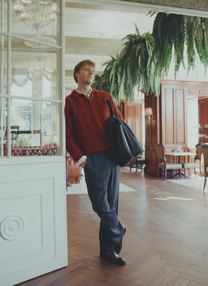 A model wearing the Percival Shugart Knit Jumper in burgundy, styled with dark trousers and a black bag indoors.
