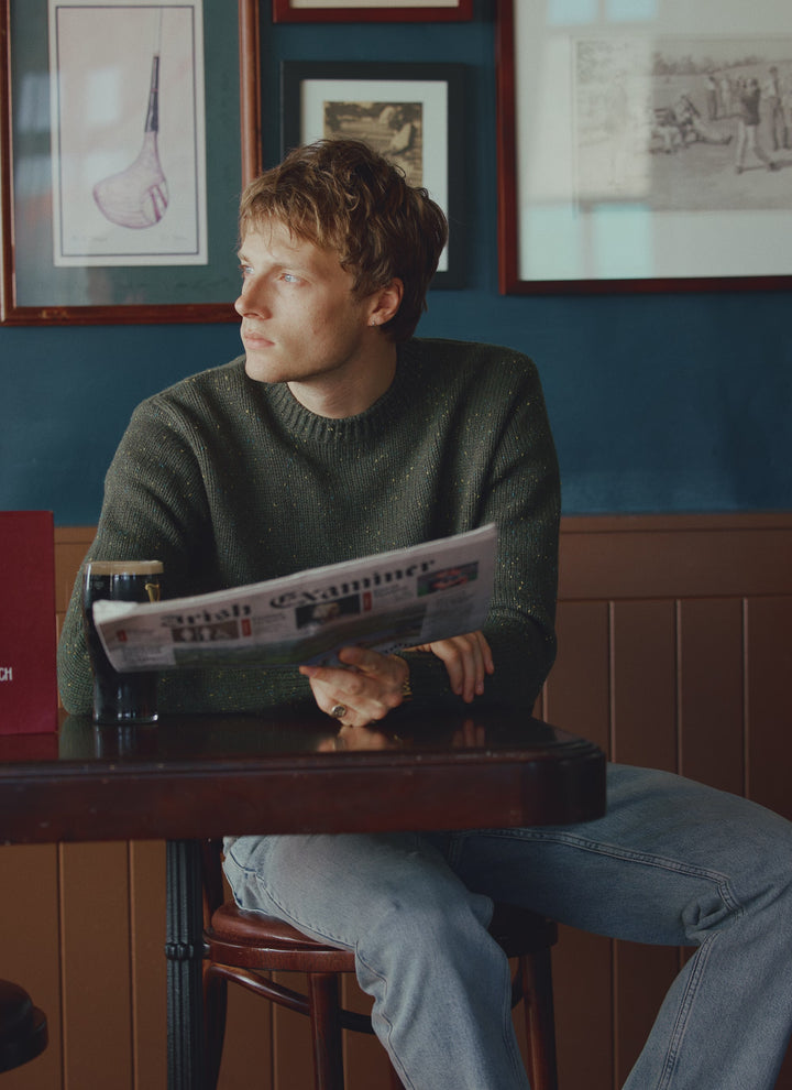 A model sits in a pub reading a newspaper, wearing the forest green Rhodes crew neck jumper from Percival.