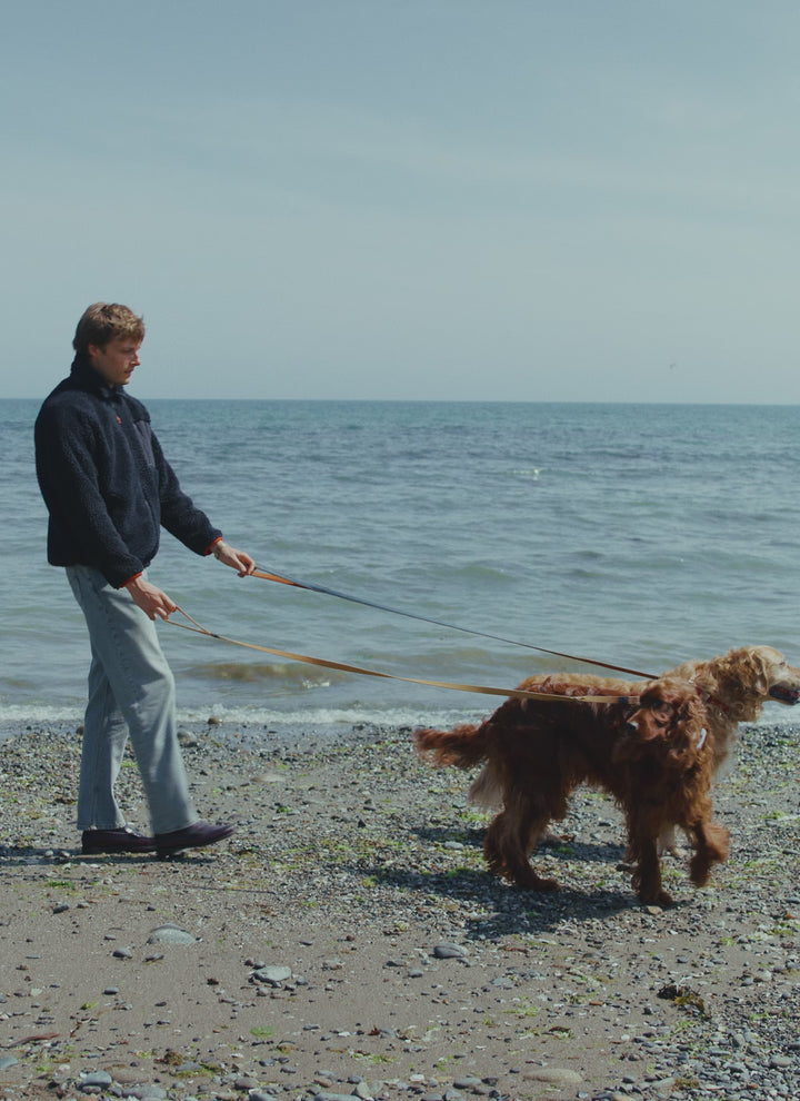 A model walks two dogs along a pebble beach while wearing a Percival navy wool blend fleece jacket and light trousers.