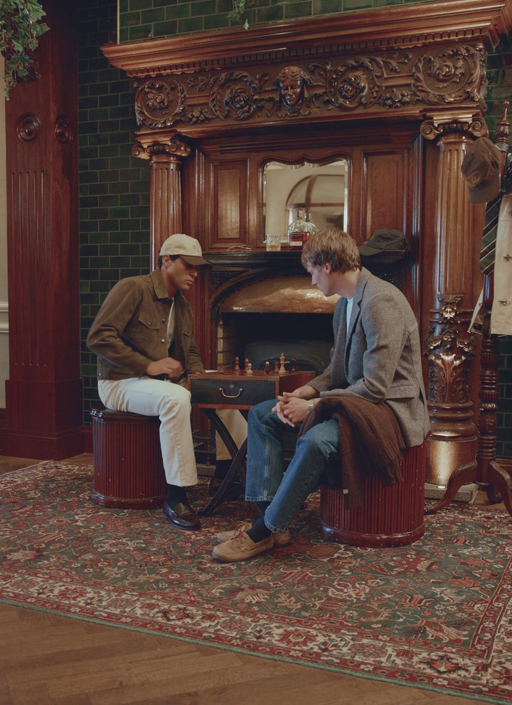 Two models playing chess in a classic interior, with one wearing the olive suede Percival Western Jacket.