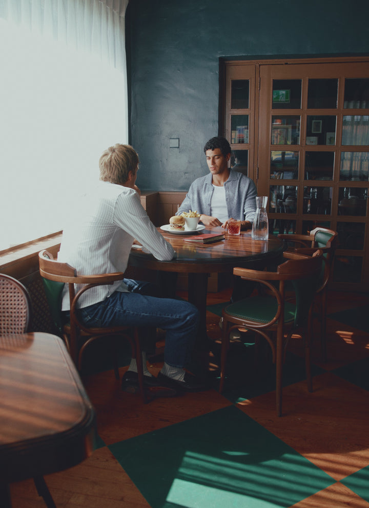 Two men sit at a restaurant table enjoying food and drinks, with one wearing the cream Percival Cuban shirt.