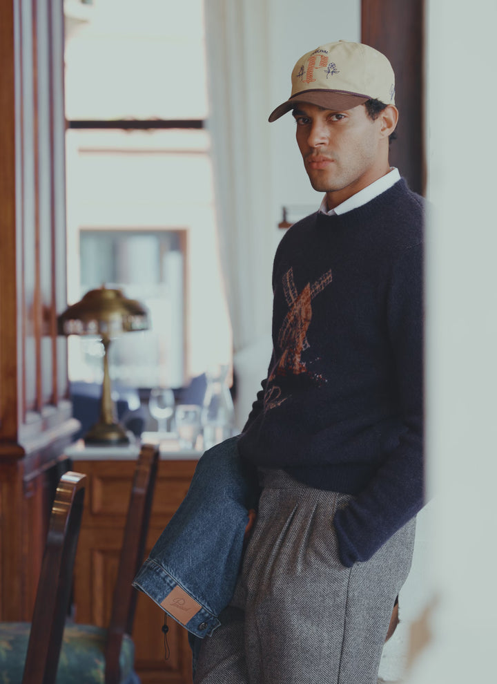 A model wearing the Percival navy Miller Jacquard Jumper featuring a windmill graphic, styled with a cap and grey trousers.