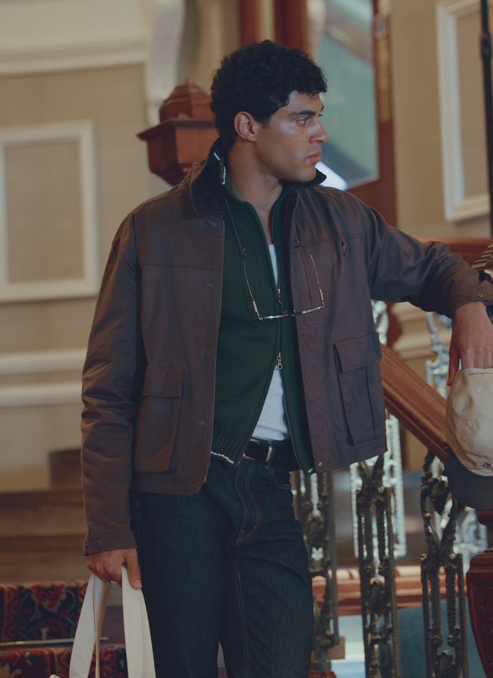 A model wears the Percival espresso brown wax zip jacket in stretch cotton, holding a tote bag indoors near a staircase.
