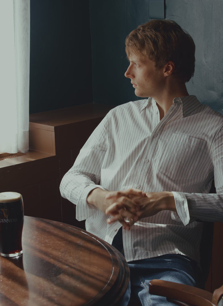 A man wears the white Percival Fabian striped Oxford shirt, sitting at a wooden table in a pub setting.